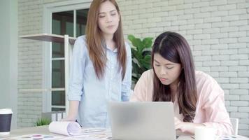 Mujeres de negocios asiáticas creativas e inteligentes atractivas en ropa casual elegante que trabajan en la computadora portátil mientras están sentadas en el escritorio en la oficina. video