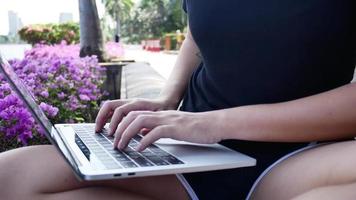 Beautiful asian young woman sitting at bench in park using laptop. video