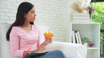 ung asiatisk attraktiv kvinna som läser intressant bok och dricker apelsinjuice i soffan i det mysiga vardagsrummet hemma. video