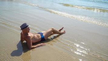 video hd stile di vita estivo di un uomo abbronzato abbastanza giovane con un cappello che si gode la vita e si siede sulla spiaggia.