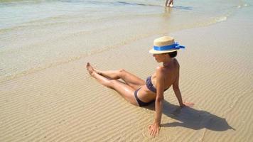 Sommerlebensstil der hübschen jungen sonnengebräunten Frau in einem Hut, der das Leben genießt und am Strand sitzt.