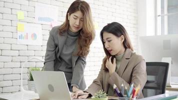 Two young Asian businesswomen working together in office at small business sitting reading a report. video