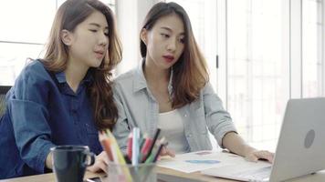 Mujeres de negocios asiáticas en ropa casual elegante que trabajan en la computadora portátil mientras están sentadas en el escritorio en la oficina. video