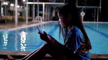 ragazza asiatica dei bambini utilizzando smart phone rilassante vicino alla piscina. video