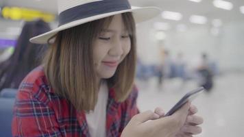 Happy Asian woman using and checking her smartphone while sitting on chair in terminal hall. video