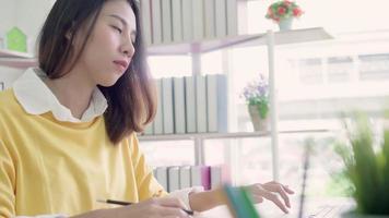 bela jovem sorridente mulher asiática trabalhando laptop na mesa na sala de estar em casa. video