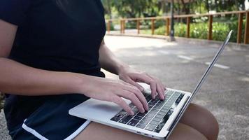 Beautiful asian young woman sitting at bench in park using laptop. video