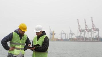 dois trabalhadores portuários de capacetes examinam documentos.