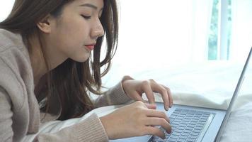 belle jeune femme asiatique souriante venant sur ordinateur portable alors qu'il était assis sur le lit dans la chambre à la maison. video