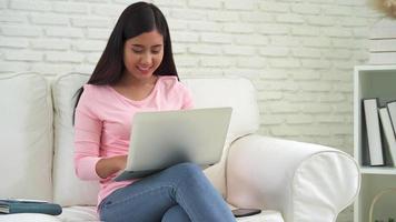 mooie Aziatische vrouw met behulp van computer of laptop liggend op de bank in haar woonkamer. video