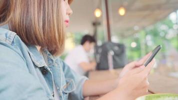Asiatische frau, die Smartphone für das Sprechen, Lesen und SMS während des Sitzens auf Tisch im Café verwendet. video