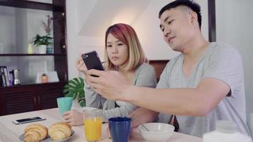 Attractive young Asian couple distracted at table with newspaper and cell phone while eating breakfast. video