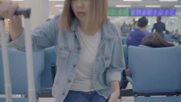 feliz mulher asiática usando e verificando seu smartphone enquanto está sentado na cadeira no corredor do terminal enquanto espera seu voo no portão de embarque no aeroporto internacional. video