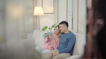 Attractive Asian sweet couple enjoy love moment drinking warm cup of coffee or tea in their hands on sofa in the living room at home. video