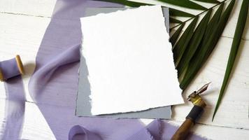 White blank greeting card ribbon on a background of blue fabric with palm leaf and calligraphic pen on a white background. Mockup with envelope and blank card. Flat lay. Top view video