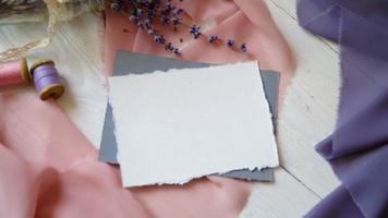 cartão em branco branco, envelope e fita em um fundo de tecido rosa e azul com flores de lavanda em um fundo branco.