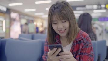 Feliz mujer asiática usando y revisando su teléfono inteligente mientras está sentado en una silla en el pasillo de la terminal.