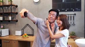 Happy young Asian couple using smartphone for selfie while cooking in the kitchen at home.