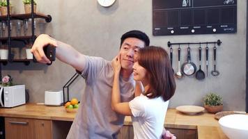 Feliz pareja asiática joven con smartphone para selfie mientras cocina en la cocina de casa. video