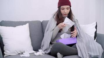 Mujer asiática joven enferma siente dolor de cabeza sentada en una silla envuelta en una manta gris en su sala de estar en casa. video