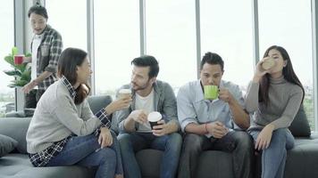 mångfald av ungdomars grupplag som håller kaffekoppar och diskuterar något med leende när man sitter i soffan på kontoret. video