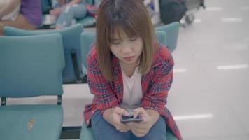 Feliz mujer asiática usando y revisando su teléfono inteligente mientras está sentada en una silla en el pasillo de la terminal mientras espera su vuelo en la puerta de salida del aeropuerto internacional. video