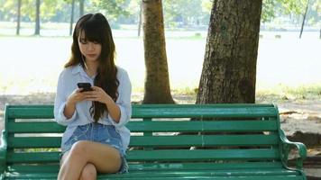 Linda mujer está leyendo un agradable mensaje de texto en el teléfono móvil mientras está sentado en el parque en un cálido día de primavera video