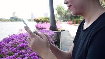 schöne asiatische junge Frau, die an Bank im Park mit Smartphone zum Sprechen, Lesen und SMS sitzt. video