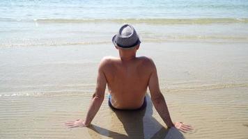 Sommer Lebensstil HD Video von hübschen jungen sonnengebräunten Mann in einem Hut genießen das Leben und sitzen am Strand.