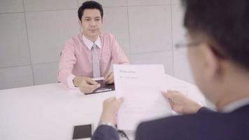 slow motion - aantrekkelijke jonge Aziatische zakenman in een sollicitatiegesprek met corporate personeelsmanager die zijn cv leest. Azië zakenman praten met mannelijke kandidaat aan bureau, sollicitant interviewen. video