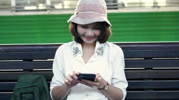 viajero, mochilero, mujer asiática, viaje, en, bangkok, tailandia. feliz mujer joven sentada en un banco con smartphone para hablar, leer y enviar mensajes de texto en la estación de tren. video