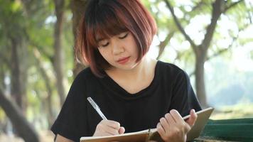 glückliche junge asiatische Frau des Hipsters, die in ihr Tagebuch im Park schreibt. video