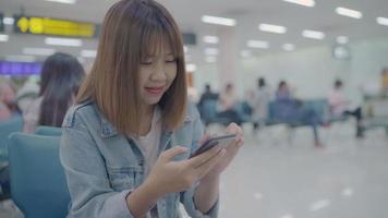 feliz mulher asiática usando e verificando seu smartphone enquanto está sentado na cadeira no corredor do terminal enquanto espera seu voo no portão de embarque no aeroporto internacional. video