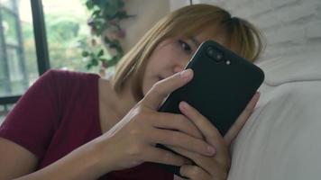 Asian woman playing smartphone while lying on home sofa in her living room. Happy female use phone for texting, reading, messaging and buying online at home. video