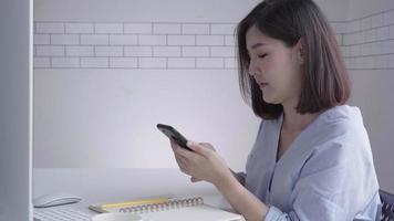 Beautiful young smiling asian woman coming on laptop while sitting in a living room at home. Asian business woman using phone for com in her home office. Enjoying time at home. video