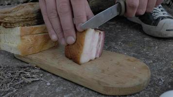 Close up of male hands slicing pork belly with knife on a wooden board outdoor. autumn picnic in the park. 4k. 4k video. slow motion. 23.98 fps video