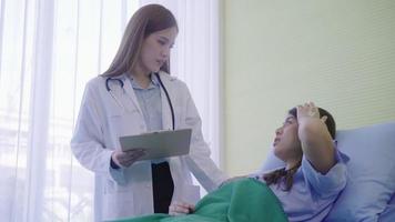 jonge Aziatische doktersvrouw toont informatiebehandeling op klembord voor vrouwelijke patiënt in ziek bed.