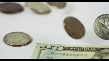  A lot of coins of quarter dollar falling and spinning over three coins and a $20 bill on white table, finally four coins and the bill placed in the scene in 4K video