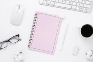 vista superior de la mesa de escritorio de oficina mínima con páginas en blanco de cuaderno rosa, computadora con teclado, mouse, taza de café en una mesa blanca con espacio de copia, composición de lugar de trabajo de color blanco, endecha plana foto