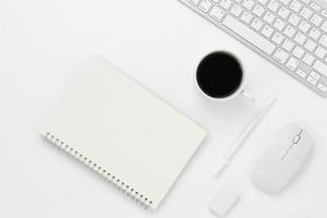 Vista superior de la mesa de escritorio de oficina mínima con páginas en blanco de un cuaderno, computadora con teclado, mouse, taza de café en una mesa blanca con espacio de copia, composición de lugar de trabajo de color blanco, endecha plana foto
