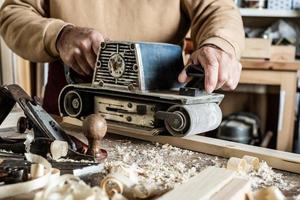 Electric belt sander, sanding machine in male hand. Processing of workpiece on light brown wooden table. Side view, close up photo