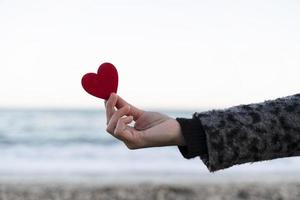 mano de mujer sosteniendo un corazón rojo en la orilla del mar. concepto de dia de san valentin foto