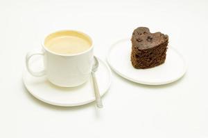 Una taza de café con leche y una cuchara junto a un pastel de chocolate sobre un fondo blanco. foto