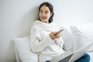 Una mujer vestida con una camisa blanca jugando con un teléfono inteligente en su cama foto