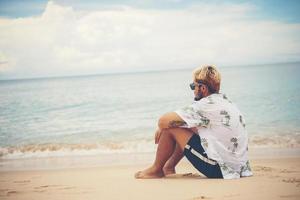 Hombre joven inconformista relajante sentado en la playa del mar foto