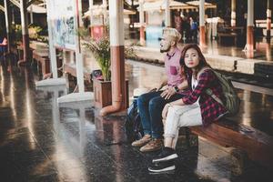 Pareja joven inconformista sentado en un banco de madera en la estación de tren foto