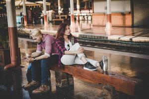 Pareja joven inconformista sentado en un banco de madera en la estación de tren foto