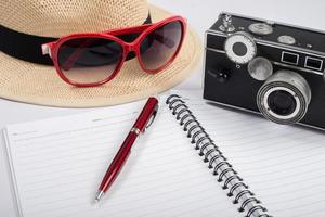 Sombrero, gafas de sol y cámara vintage con bloc de notas aislado sobre un fondo blanco. foto