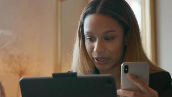 Joven empresaria negra, sosteniendo el teléfono celular frente a ella, hablando, mirando el teléfono celular y la computadora portátil video