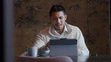 Joven asiático sentado a la mesa con un portátil, conversando por videollamada video